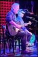 A person playing guitar and sitting on a stool

Description automatically generated