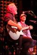 A person playing a guitar with another person in the background

Description automatically generated
